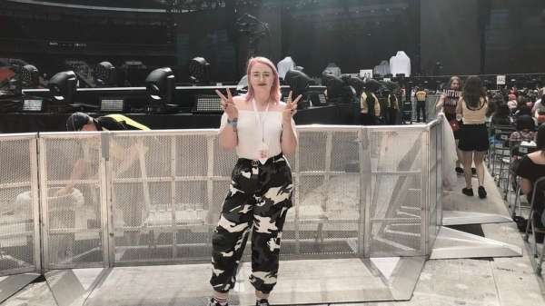Beth is posing for a photo at a K-Pop BTS concert. She has a pink bob and is posing with her fingers held up in two v shapes. She's wearing a white top and black and white cargo pants. She is stood in front of a metal gate that is shoulder height. In the background you can see all the concert stage, stewards and lights.